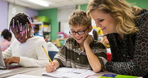 young girl with beads in hair typing at computer sitting next to young boy with glasses writing on paper with teacher leaning over next to him