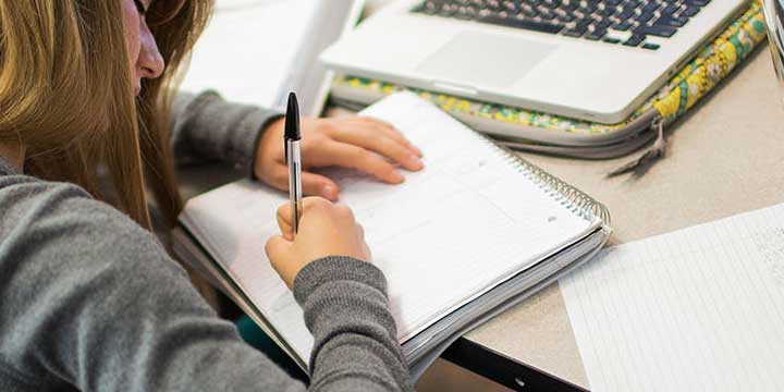 Young woman writing in spiral notebook on desk infront of laptop